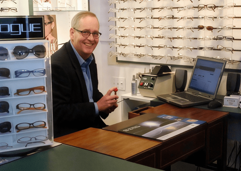 Man sitting at a desk in front of a display of glasses, and holding an object in his hand